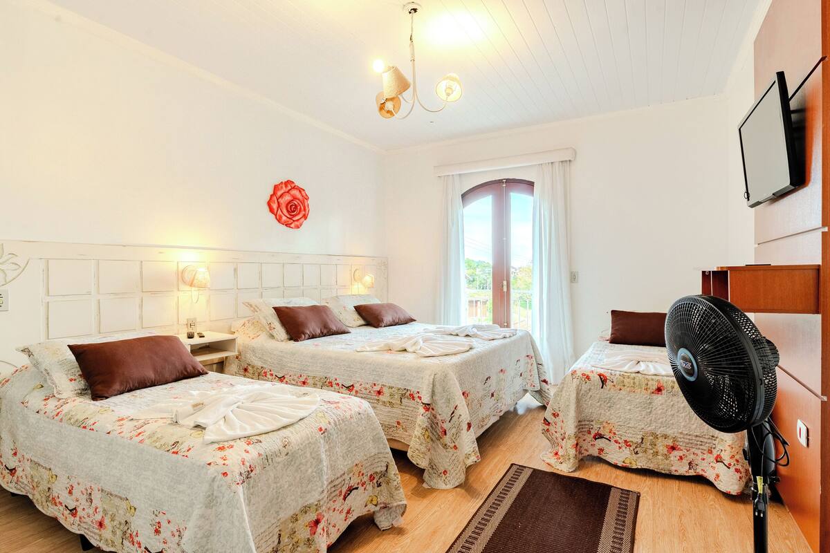 Quarto com duas camas, um ventilador de teto, janela com cortinas e televisão na parede.