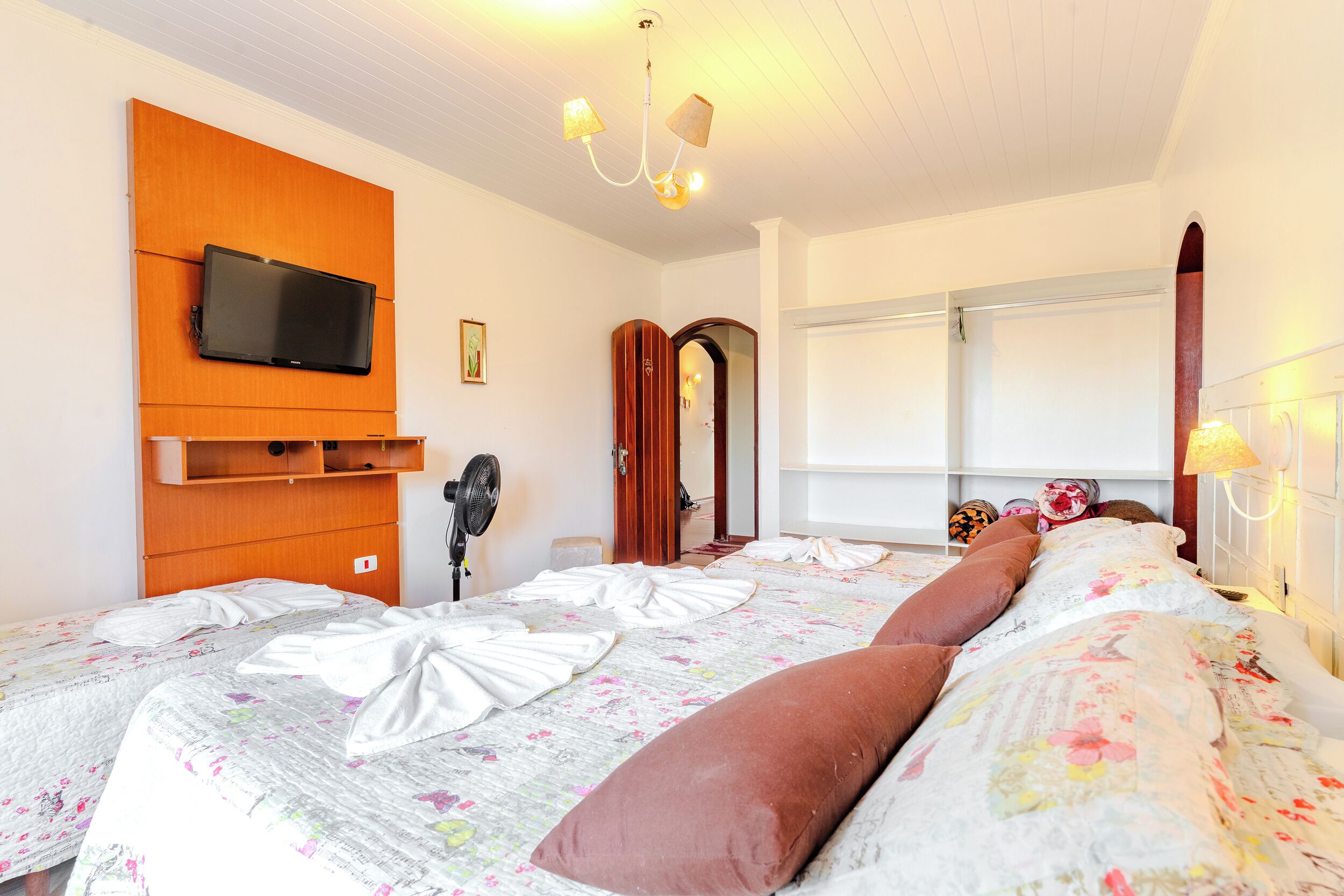 A hotel room with a large bed, a television mounted on a wooden panel, a ceiling fan, and a window with blinds.
