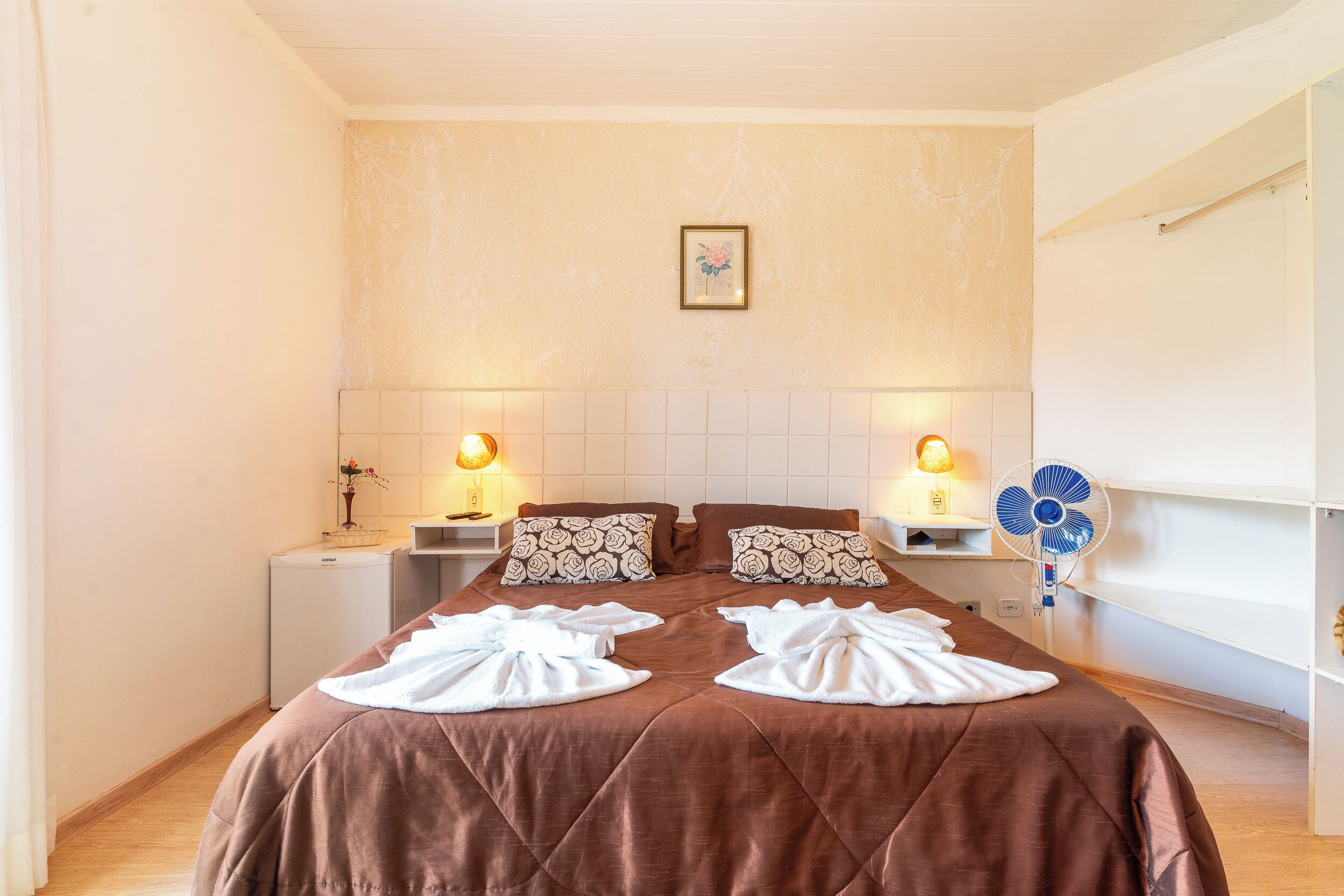 A hotel room with a large bed, two bedside tables, a wall-mounted fan, and a small shelf with decorative items.