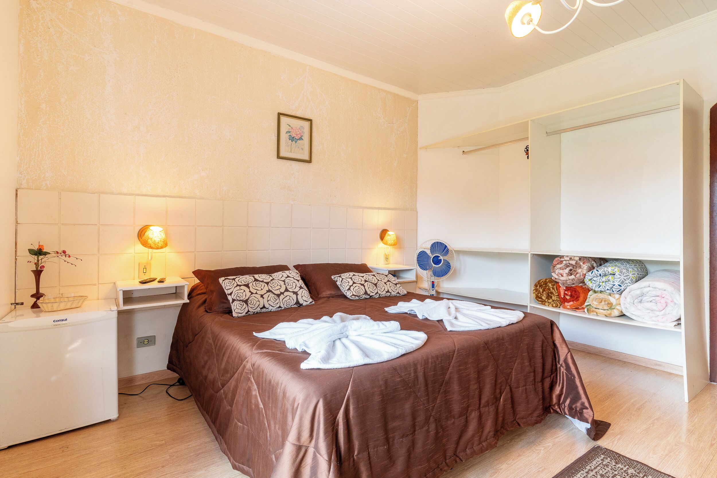 A hotel room with a bed, bedside tables, a fan, a mirror, and a shelf with towels.