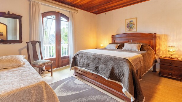A bedroom with a wooden ceiling, a large bed with a headboard, a chair, a nightstand, and a window with curtains. - Pousada Recanto Sião (Campos do Jordão)