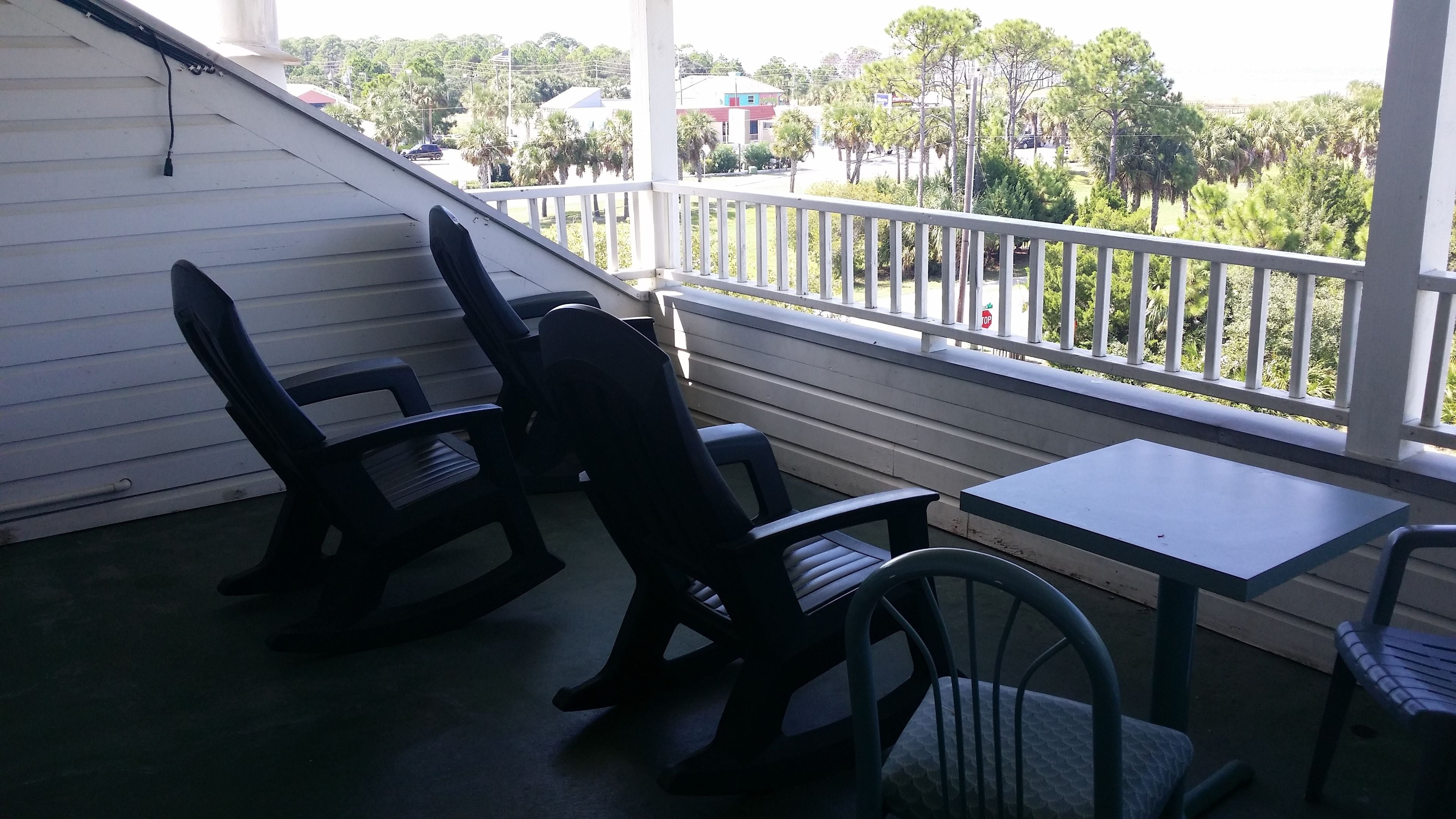 A covered outdoor seating area with two rocking chairs and a table, overlooking a street with palm trees and buildings.