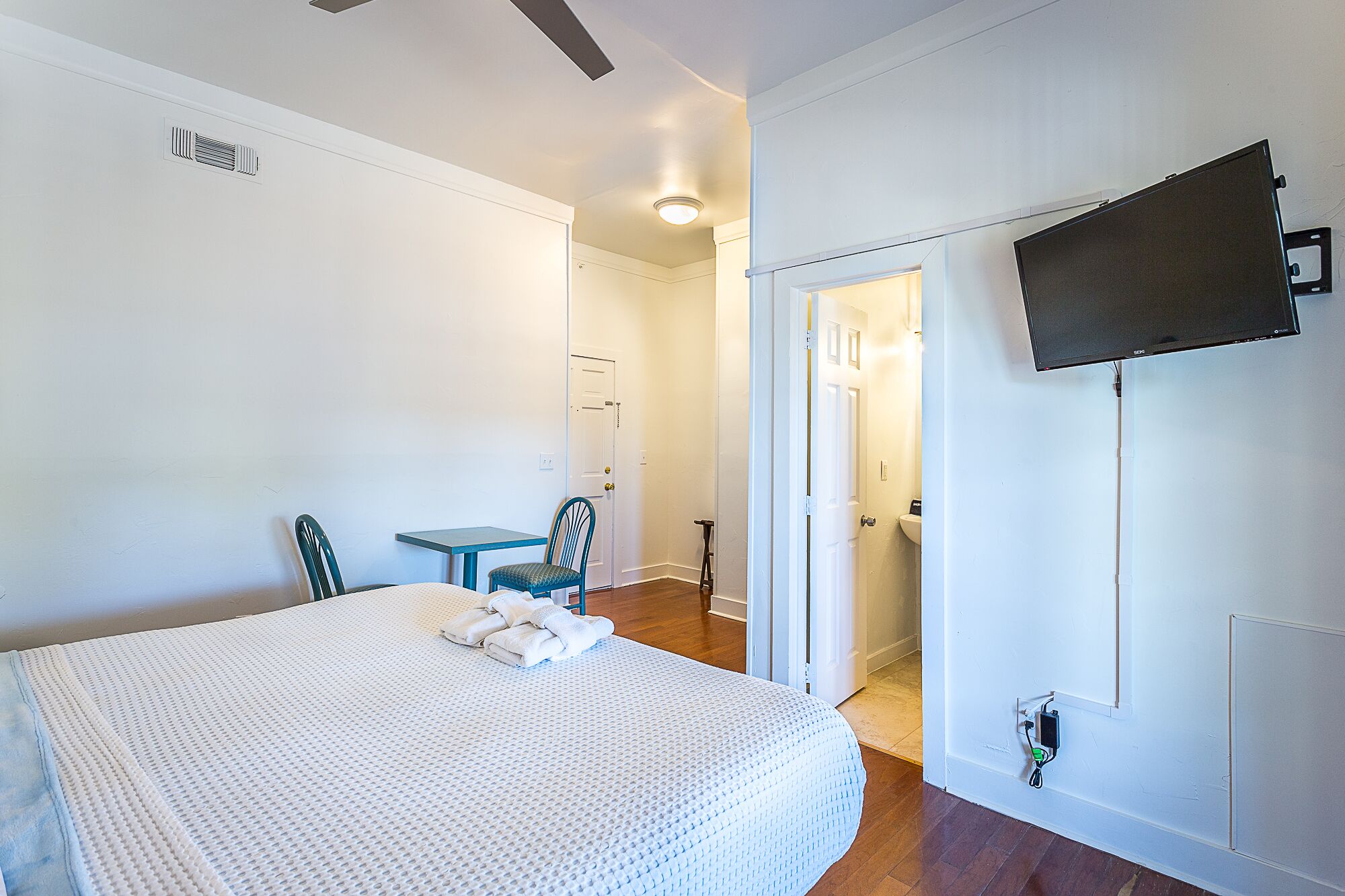 Habitación de hotel con cama, una mesita, dos sillas, televisor montado en la pared y una puerta que da a un baño.