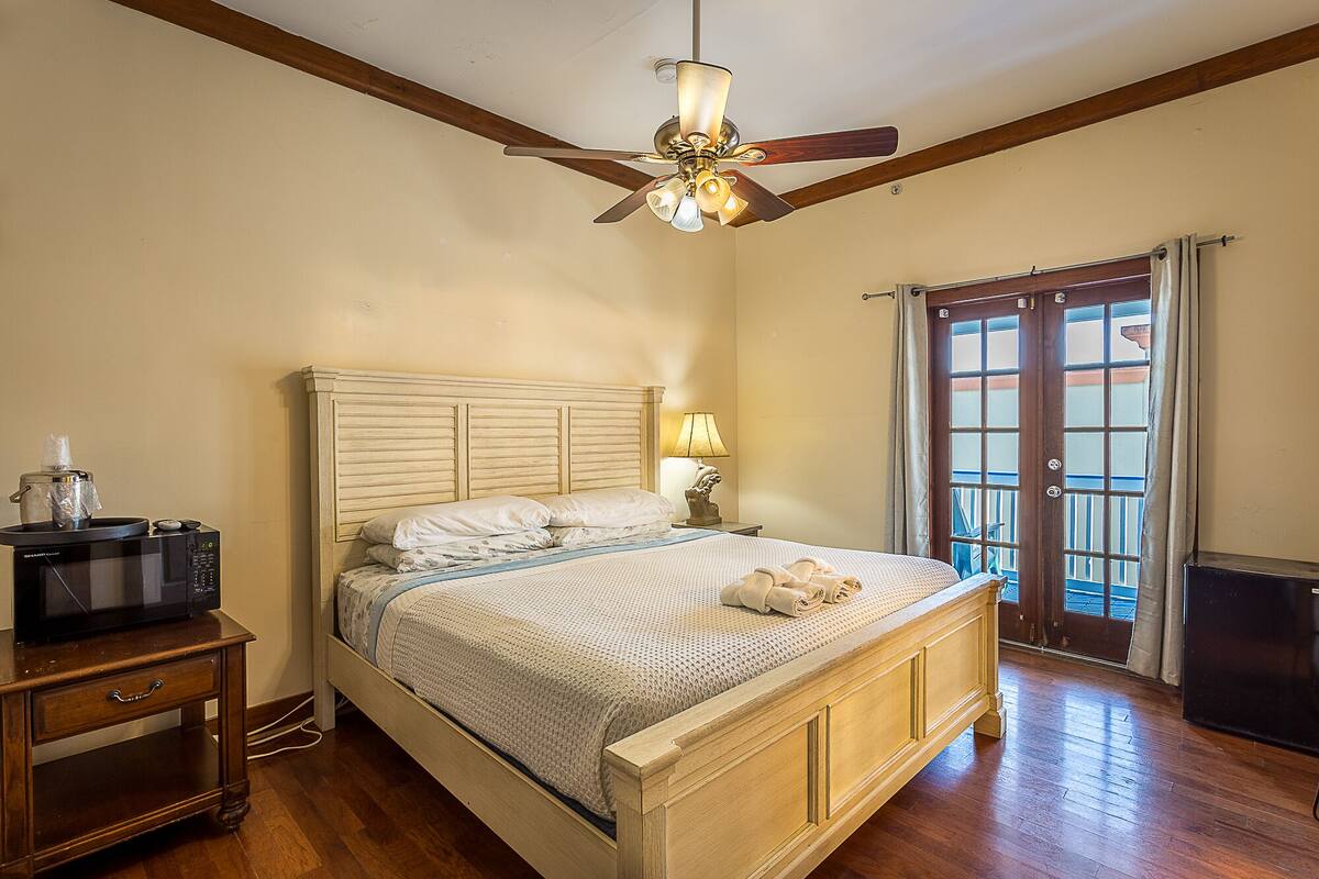 Une chambre avec un lit en bois, un ventilateur de plafond, une table de chevet, une lampe et une vue sur l’extérieur grâce à une porte vitrée.