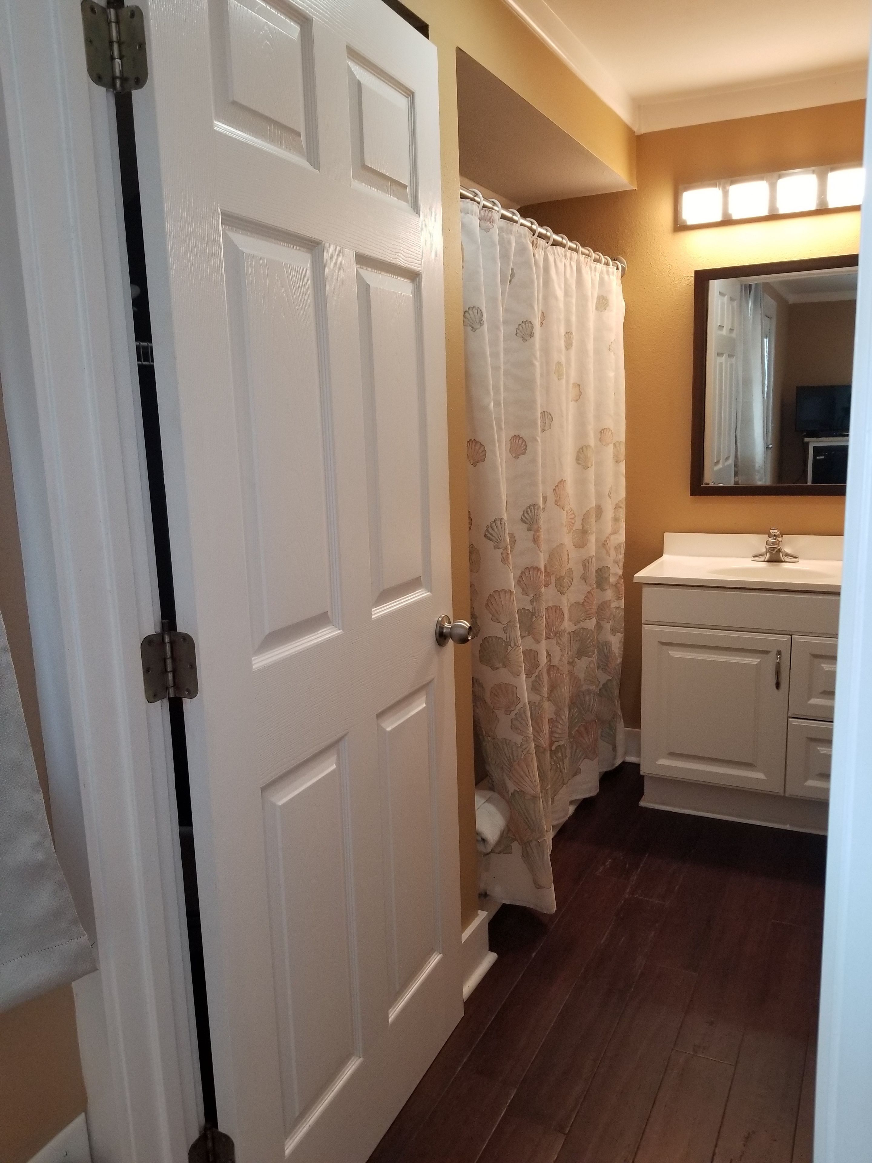 Une salle de bain avec une porte blanche, un rideau de douche orné de motifs de coquillages, un lavabo avec un miroir au-dessus et un plancher en bois.