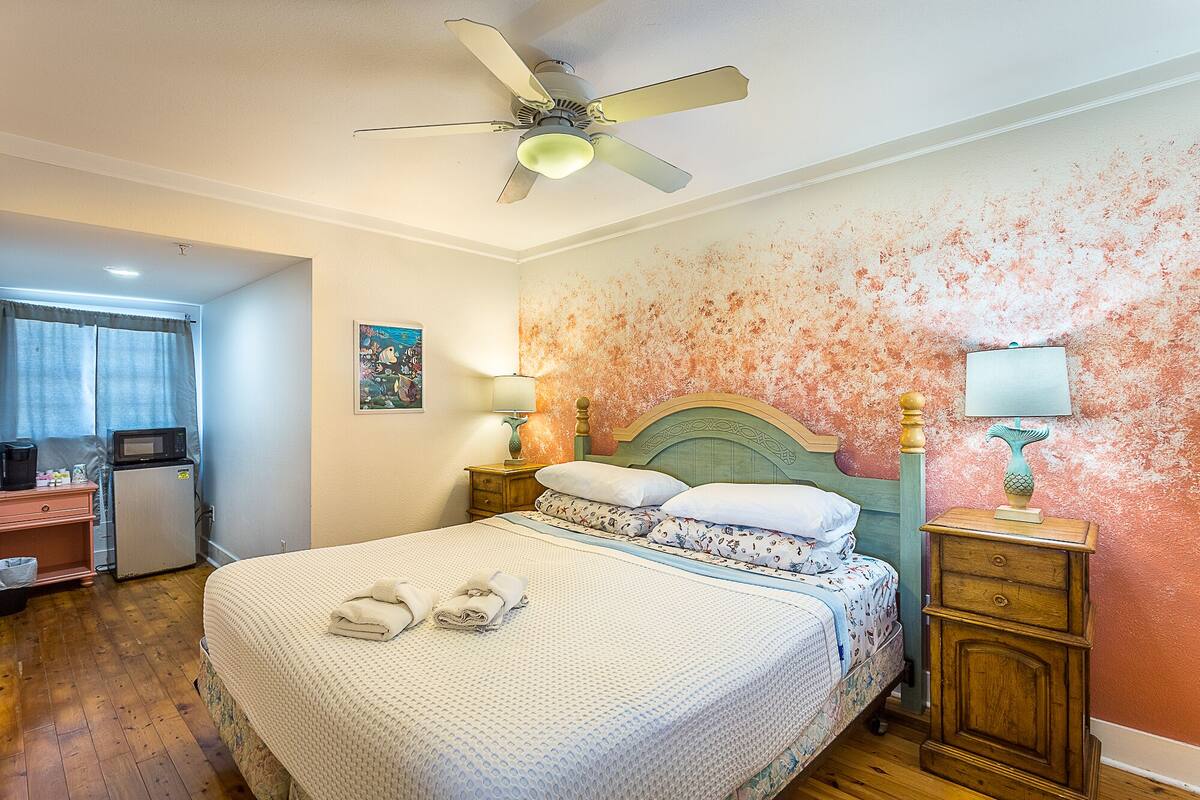 Une chambre à coucher comprenant un lit, deux tables de chevet, un ventilateur de plafond, un mur au décor texturé et une porte donnant sur une autre pièce.