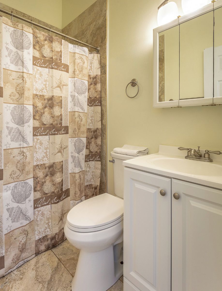 Un bagno con pavimento piastrellato color beige, un WC bianco, un lavandino bianco con rubinetto argentato e una tenda da doccia con motivo marino.
