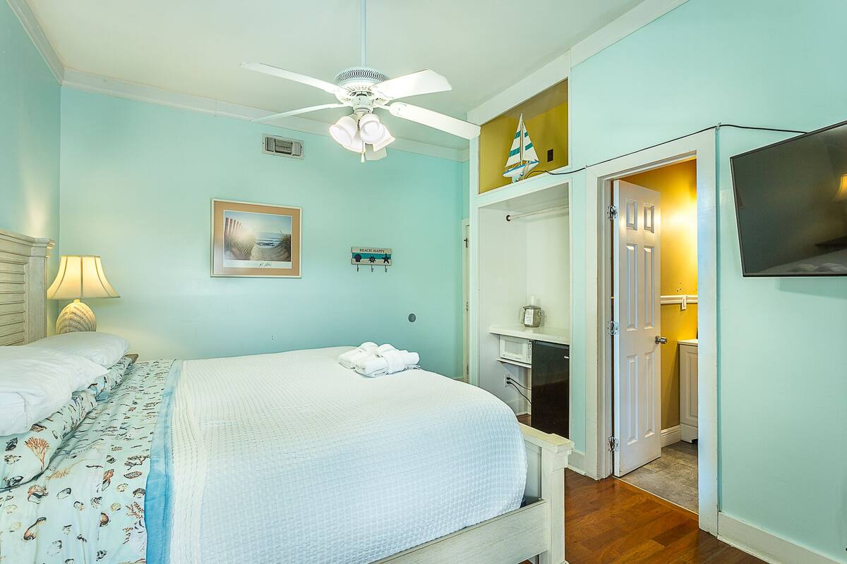 Une chambre avec un lit, un ventilateur de plafond, un tableau encadré, une pancarte sur laquelle on peut lire « PASSEZ DE BONNES JOURNÉES À LA PLAGE » et un téléviseur.