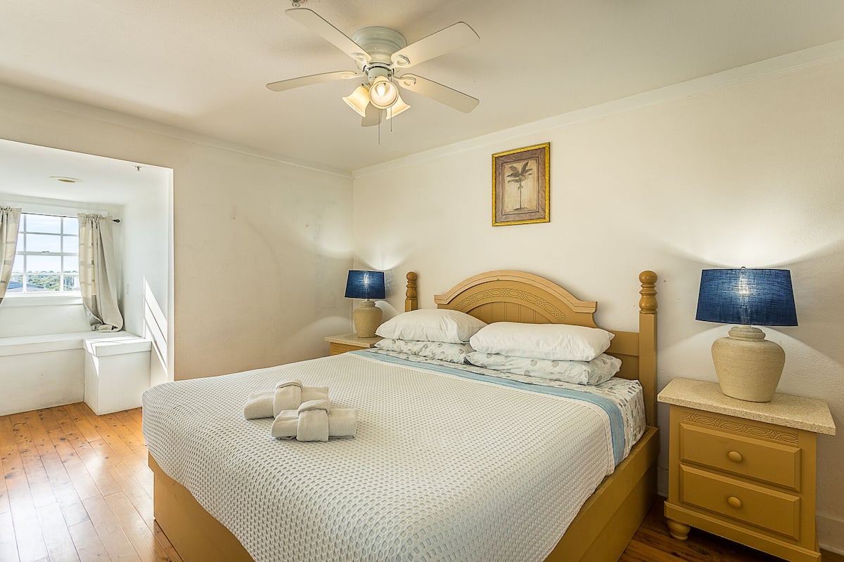 Une chambre à coucher bien rangée, avec un lit, des lampes de chevet, une table de nuit, un ventilateur de plafond et une fenêtre avec des rideaux.