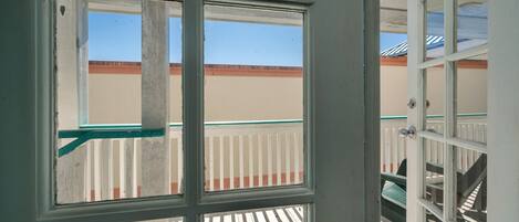 A view through a window with a white frame and green trim, looking out to a balcony with white railings and a beige wall in the background.