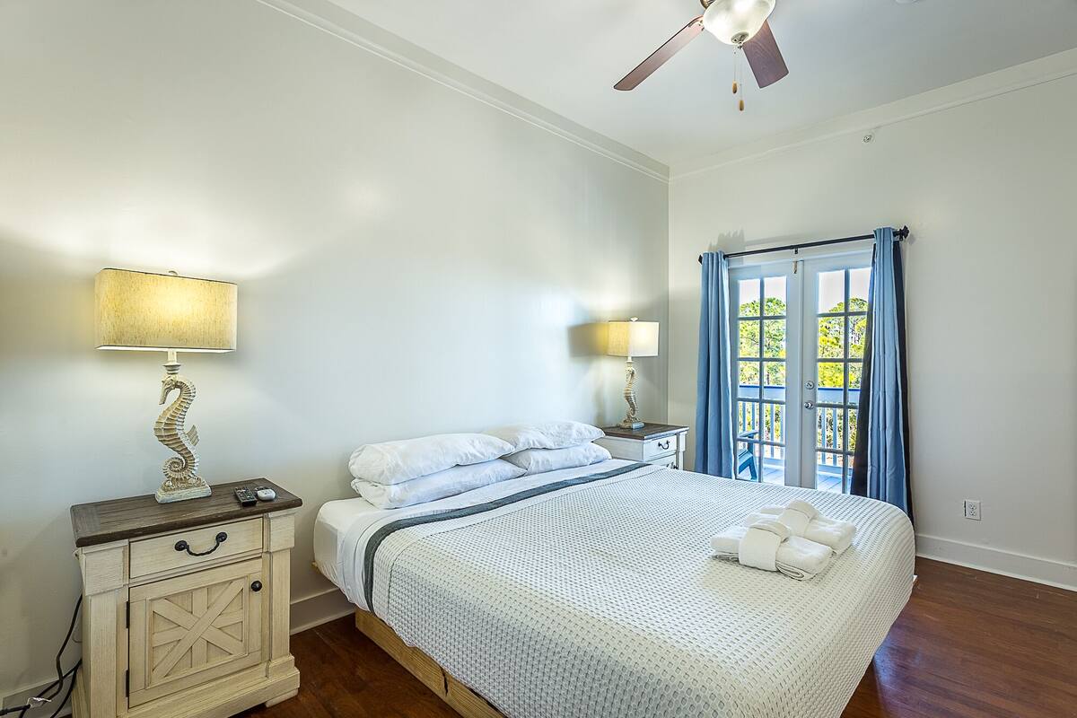 Une chambre à coucher avec un lit, deux tables de chevet avec des lampes, un ventilateur de plafond et une fenêtre avec des rideaux.