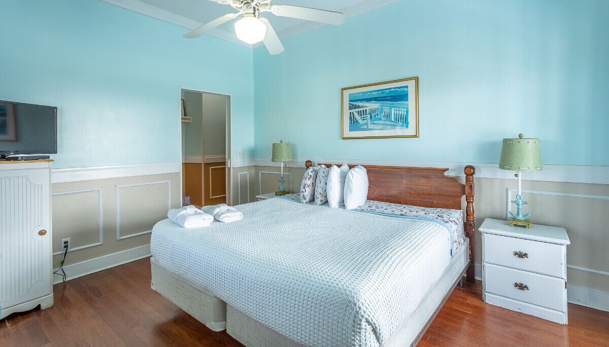 A bedroom with a bed, a nightstand, a lamp, a ceiling fan, and a painting on the wall.