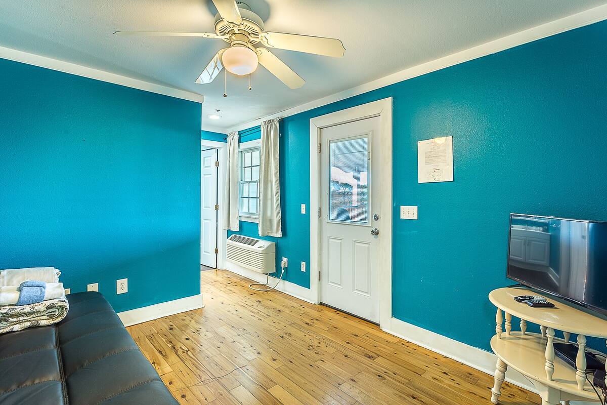 Une pièce aux murs de couleur turquoise, avec un ventilateur de plafond, un plancher en bois, une porte avec une fenêtre, une petite table sur laquelle se trouve un téléviseur et un canapé.