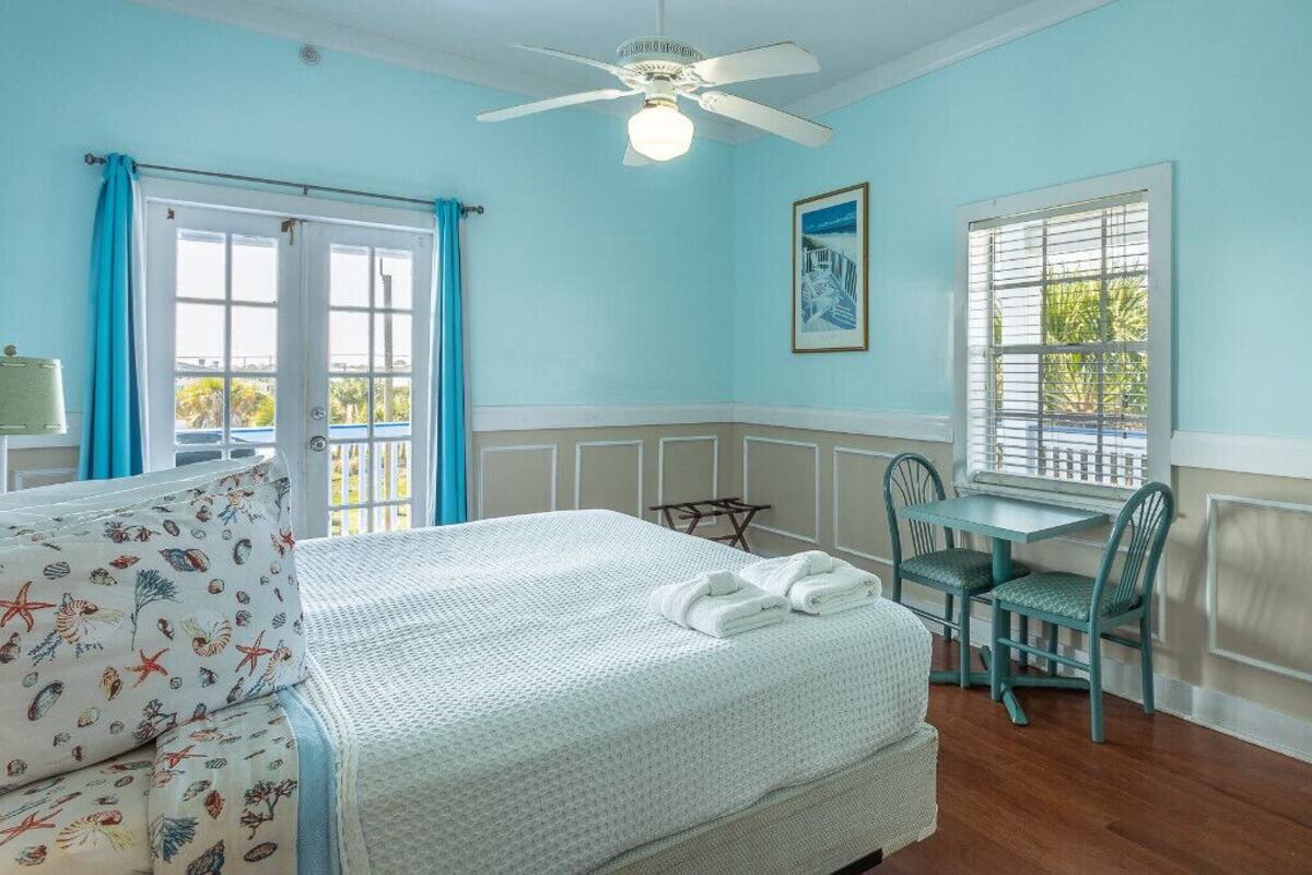 Une pièce comprenant un lit, un bureau et une chaise. On y trouve également un ventilateur de plafond, une fenêtre et un tableau encadré accroché au mur.