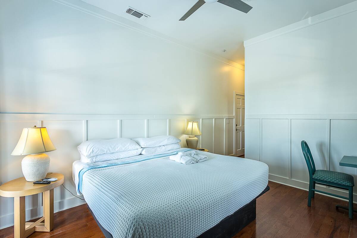 Un lit bien fait, recouvert d’une couvre-lit à motifs bleus et blancs, deux lampes de chevet, une chaise et un ventilateur de plafond.