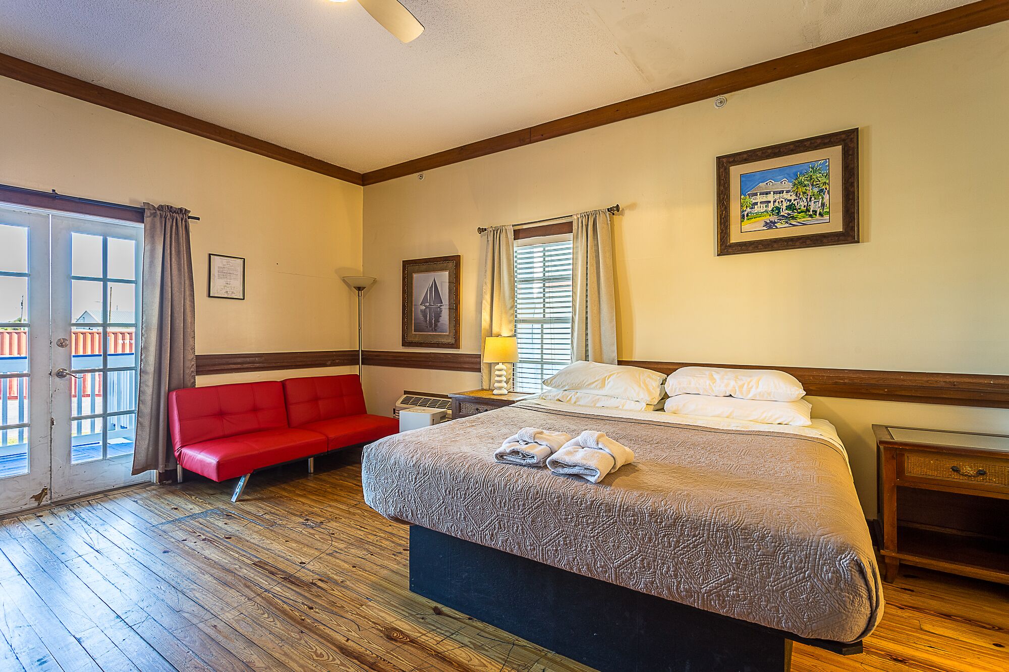 A hotel room with a large bed, a red sofa, a wooden floor, and a balcony with a view.