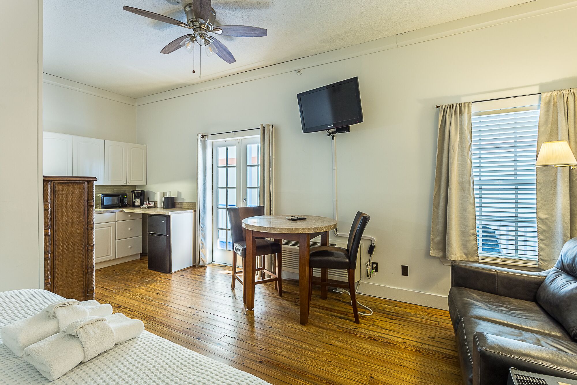A compact living space with a kitchenette, a dining area with a table and chairs, a ceiling fan, a flat-screen TV mounted on the wall, and a leather sofa.