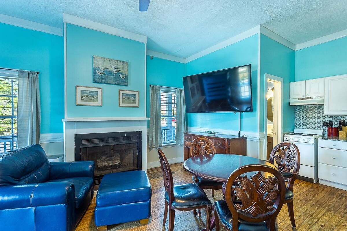 Un salon avec une cheminée, une table à manger avec des chaises, un téléviseur fixé au mur et un coin cuisine avec des placards et une cuisinière.