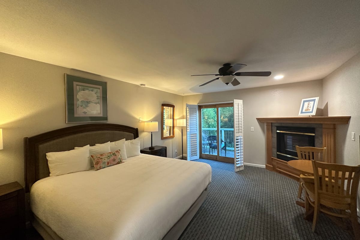 A hotel room with a large bed, a ceiling fan, a fireplace, and a view of the outdoors.
