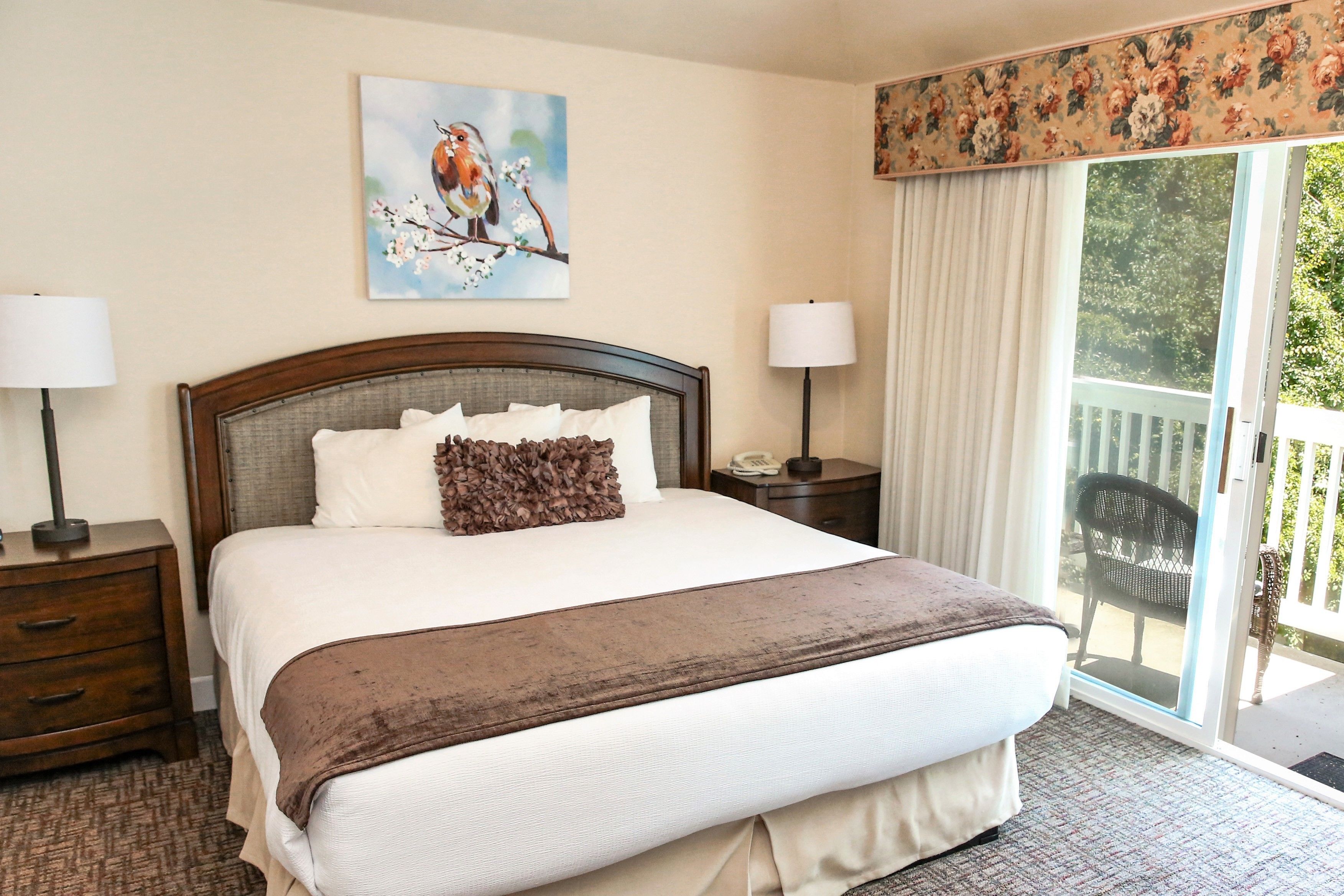 A hotel room with a large bed, two bedside tables with lamps, a painting of a bird on the wall, and a view of the outdoors through a sliding door.