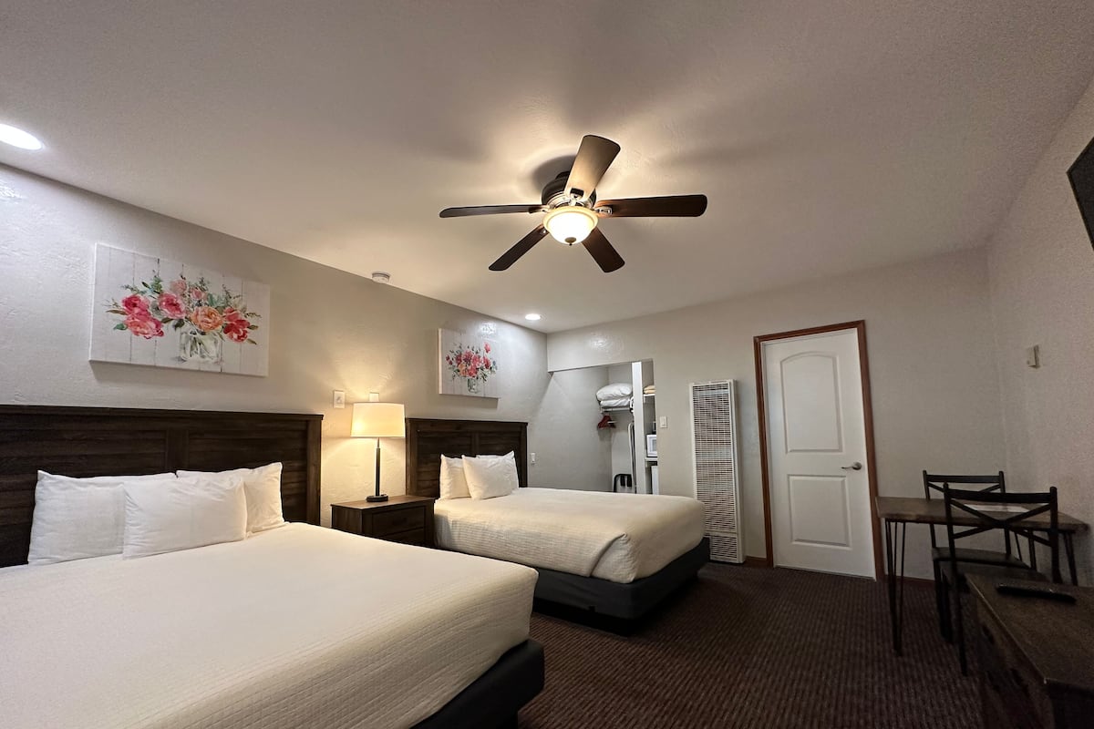 A hotel room with two beds, a ceiling fan, a desk with chairs, and a door.