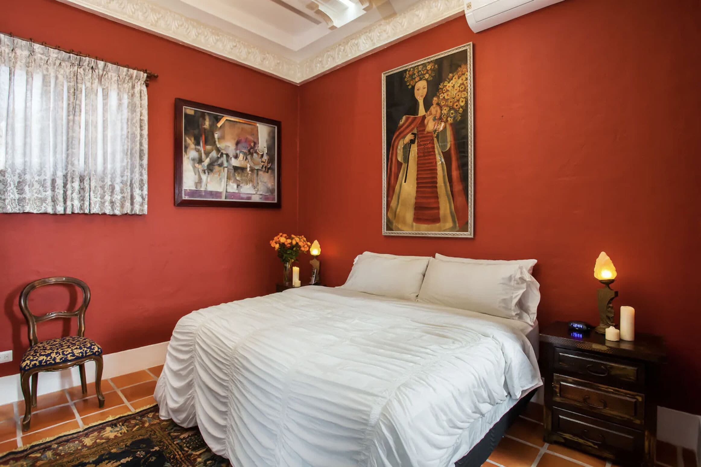 A bedroom with a large bed, a chair, a nightstand with a lamp, and two paintings on the wall.