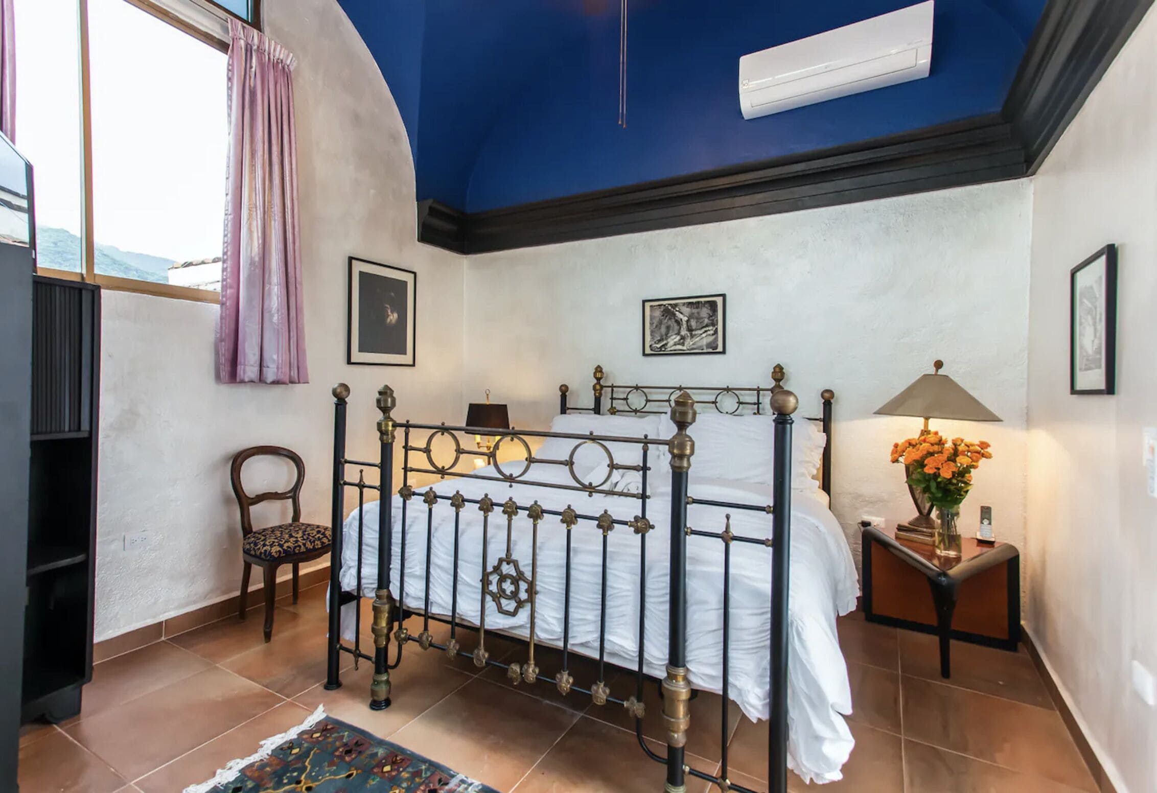 Een slaapkamer met een bed, een stoel, een nachtkastje met een lamp en een raam met gordijnen.