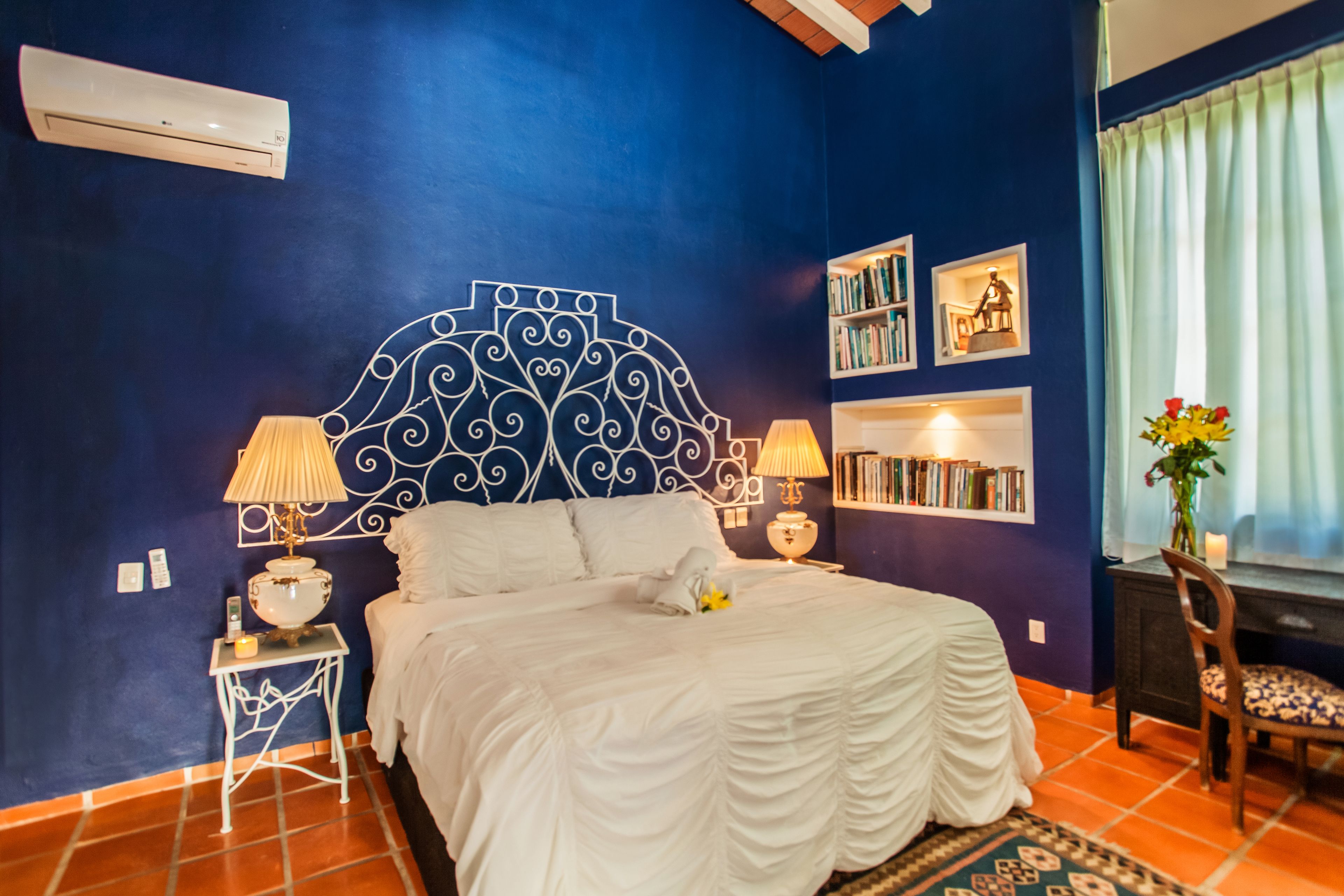 A bedroom with a blue wall, a decorative headboard, a bed with white bedding, two bedside tables with lamps, a bookshelf, and a window with curtains.