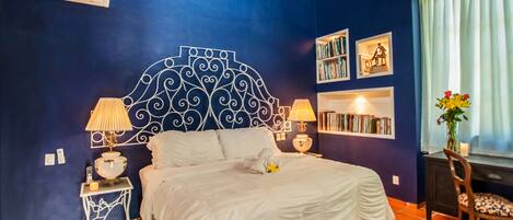 A bedroom with a blue wall, a decorative headboard, a bed with white bedding, two bedside tables with lamps, a bookshelf, and a window with curtains.