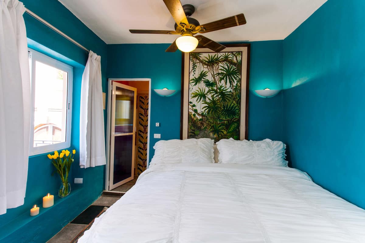 Un dormitorio con una cama, un ventilador de techo, una ventana con cortinas y una puerta que da a otra habitación.