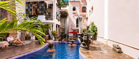 A swimming pool with a tiled edge, surrounded by buildings with balconies and potted plants.