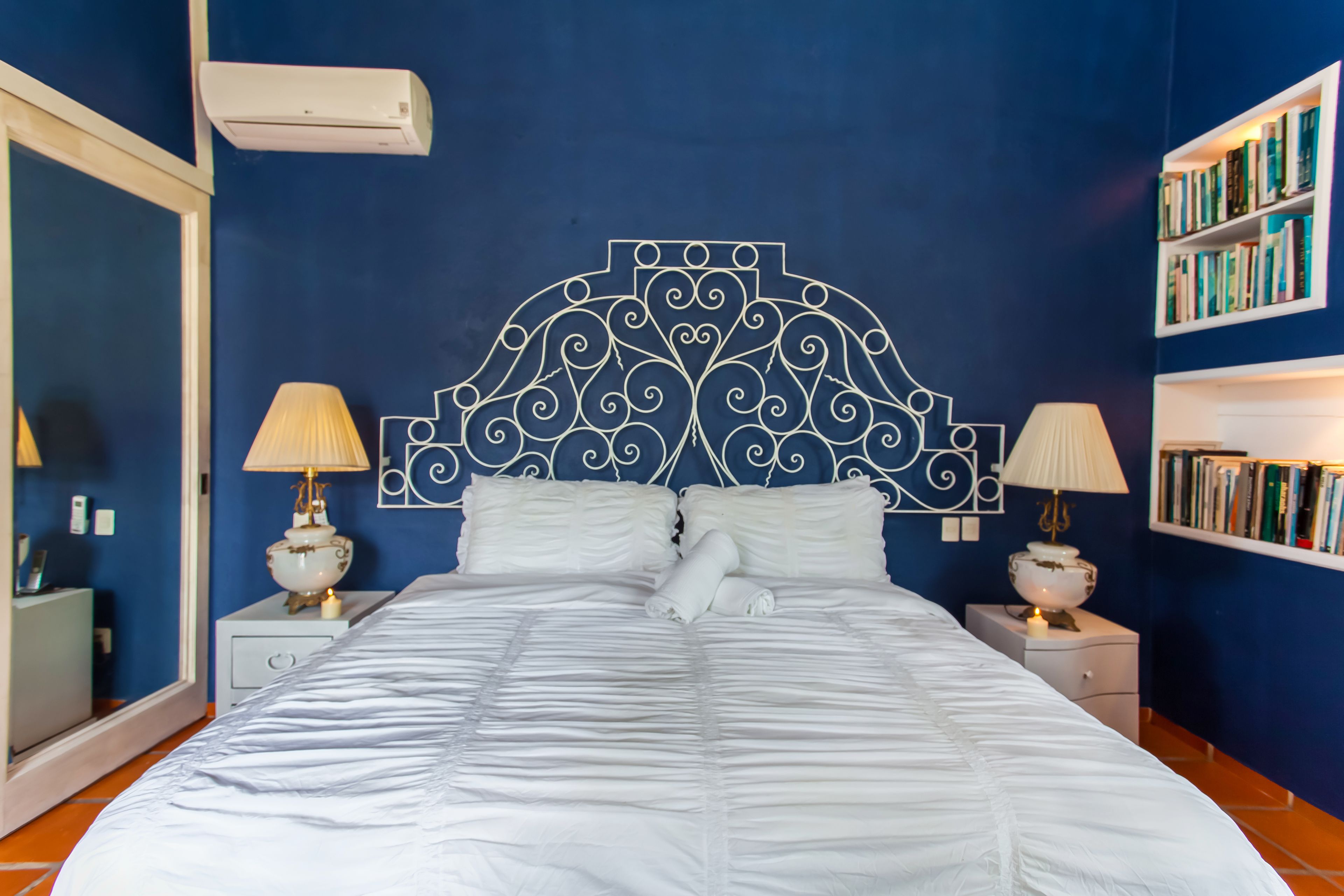 A neatly made bed with white linens, a decorative headboard, two bedside lamps, and a bookshelf with books.