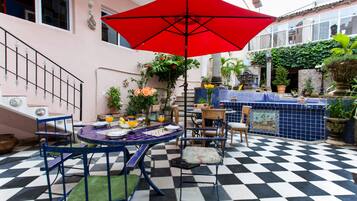 Une terrasse extérieure à carreaux avec une table et des chaises, un parasol rouge et des plantes en pot.