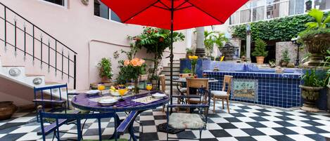 Un patio al aire libre con piso a cuadros, una mesa y sillas, un paraguas rojo y plantas en macetas.