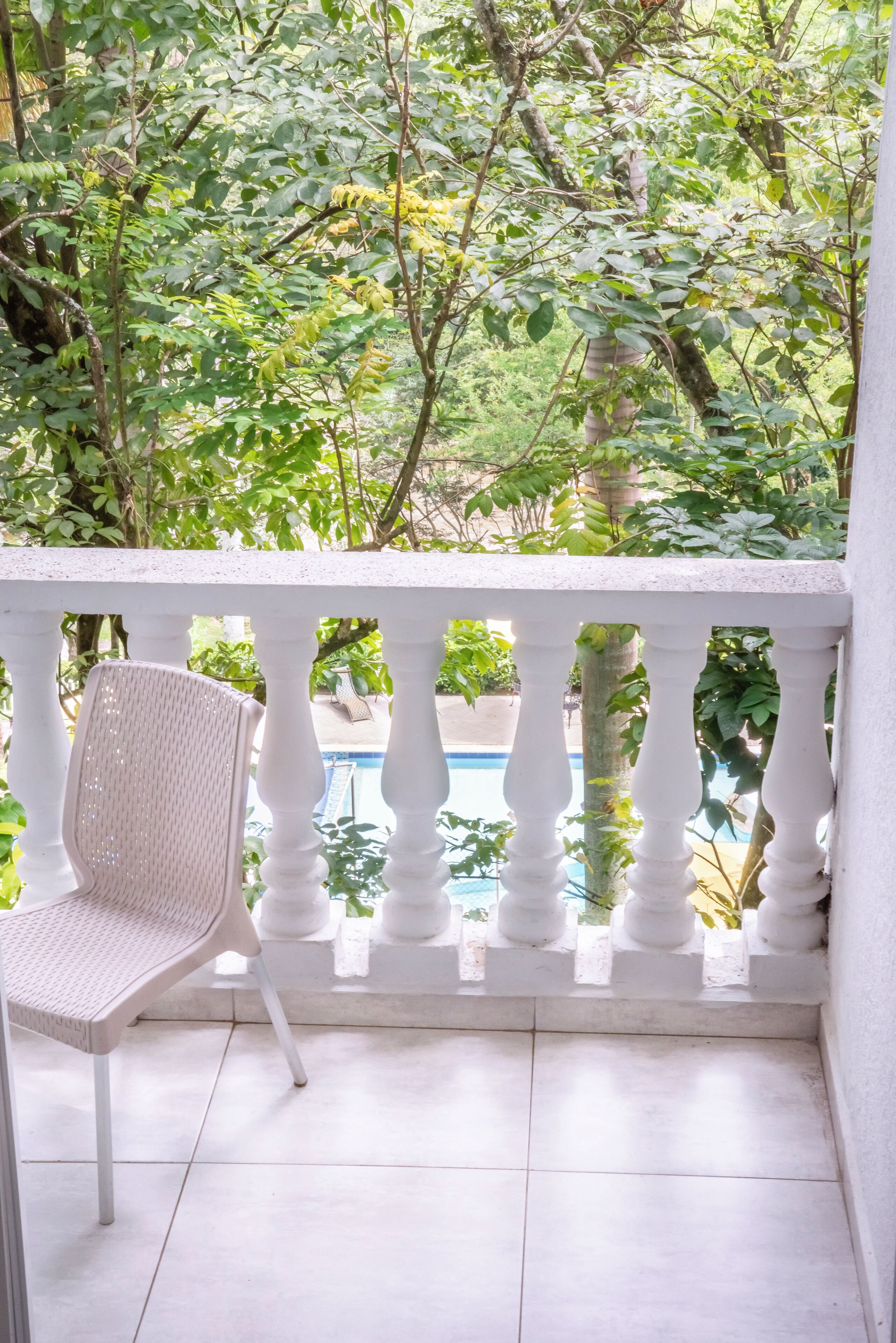 A balcony with a white chair, a pool, and lush greenery in the background.