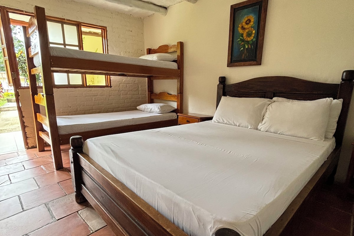 A room with a wooden bunk bed, a single bed with white bedding, a small bedside table, and a painting of sunflowers on the wall.