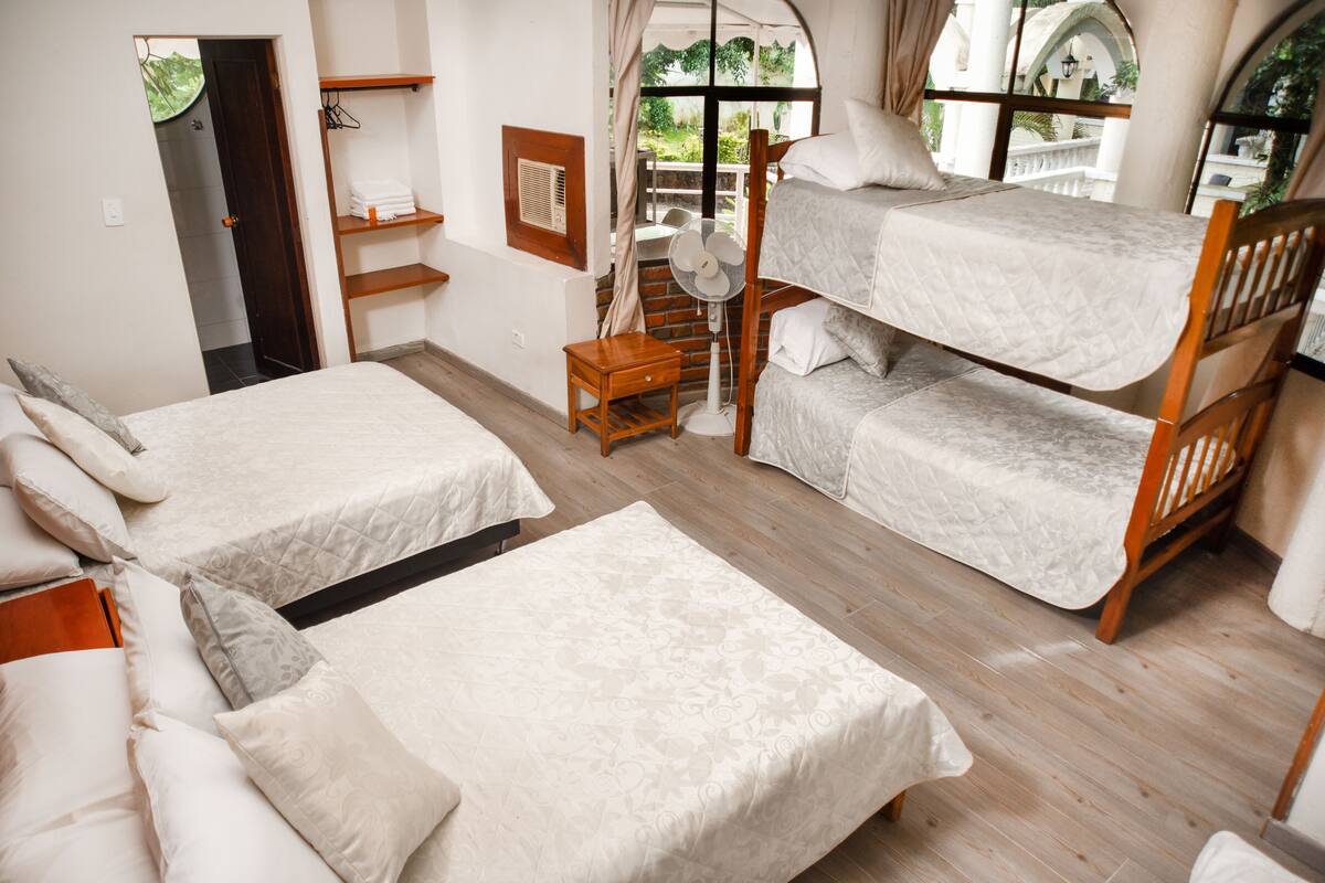 A room with two single beds and a bunk bed, a small wooden nightstand, a fan, and a window with curtains.