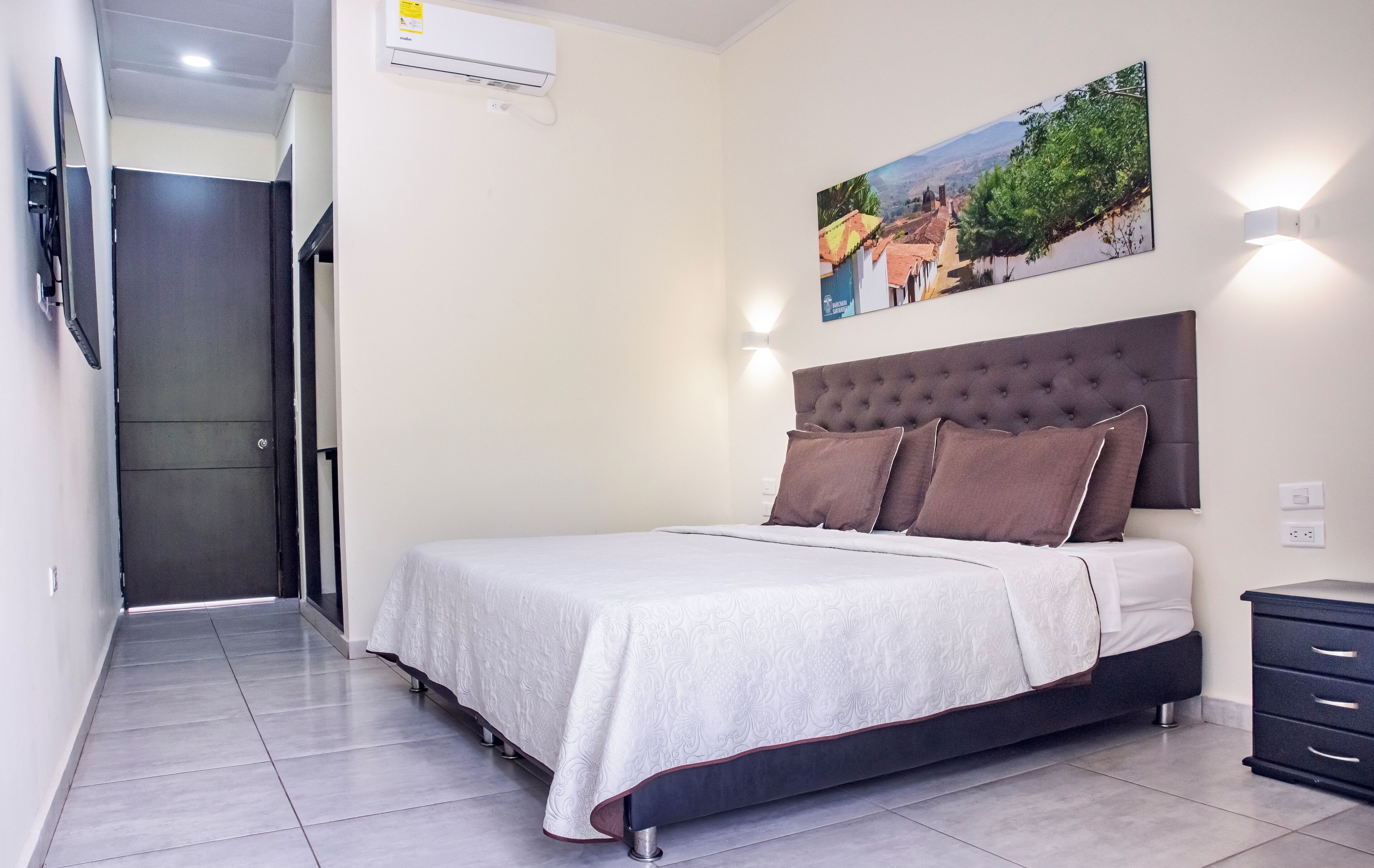 Un dormitorio con cama, cabecera, mesita de noche, televisor montado en la pared y un cuadro.