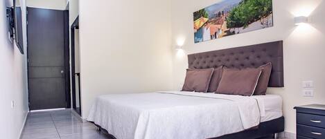 Un dormitorio con cama, cabecera, mesita de noche, televisor montado en la pared y un cuadro.