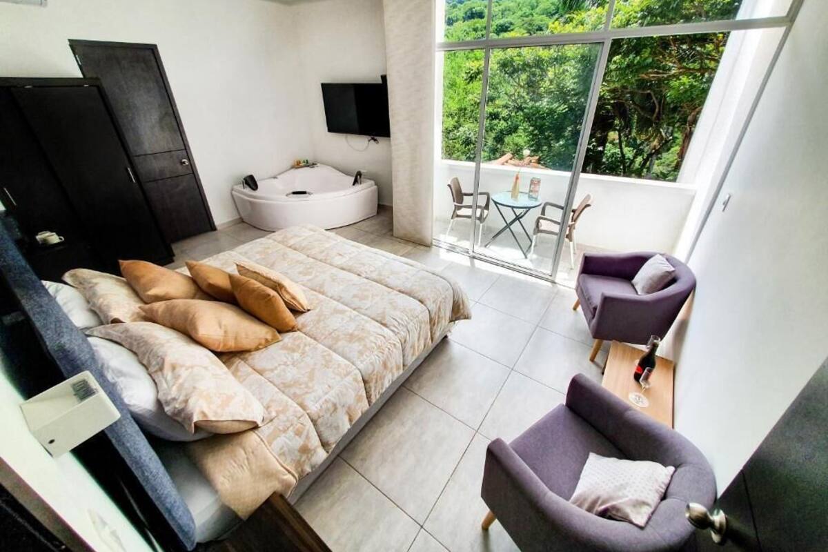 A bedroom with a large bed, a sitting area with a table and chairs, a bathtub, and a balcony with a view of greenery.