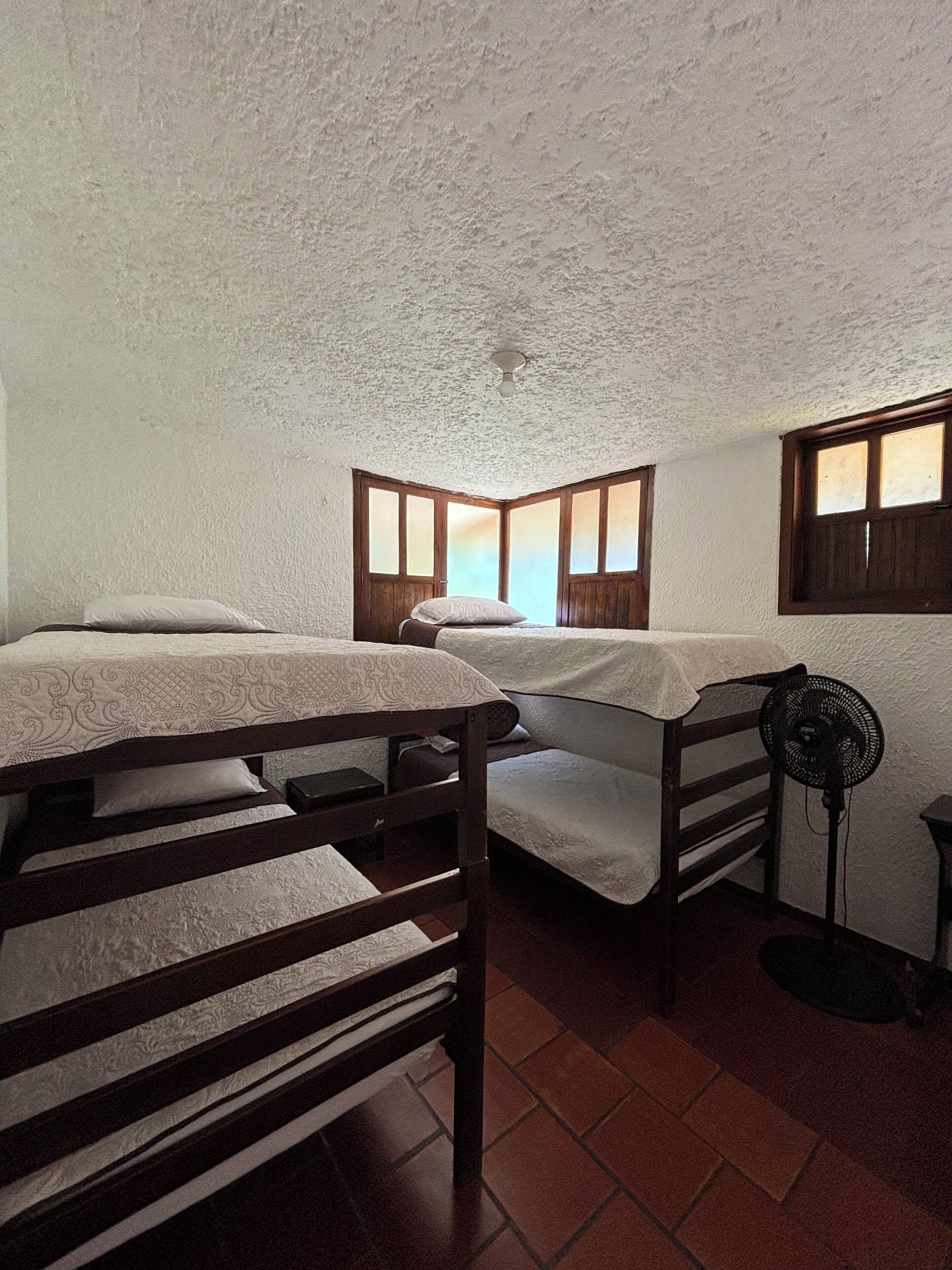 A dormitory room with two bunk beds, a ceiling fan, and a window with wooden shutters.
