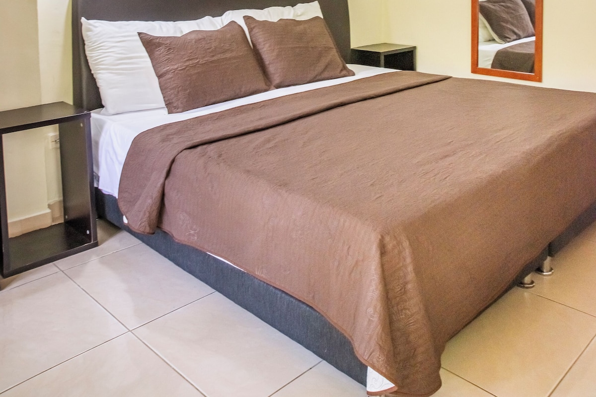 A neatly made bed with a brown bedspread, a headboard, and a painting of a forest on the wall above.