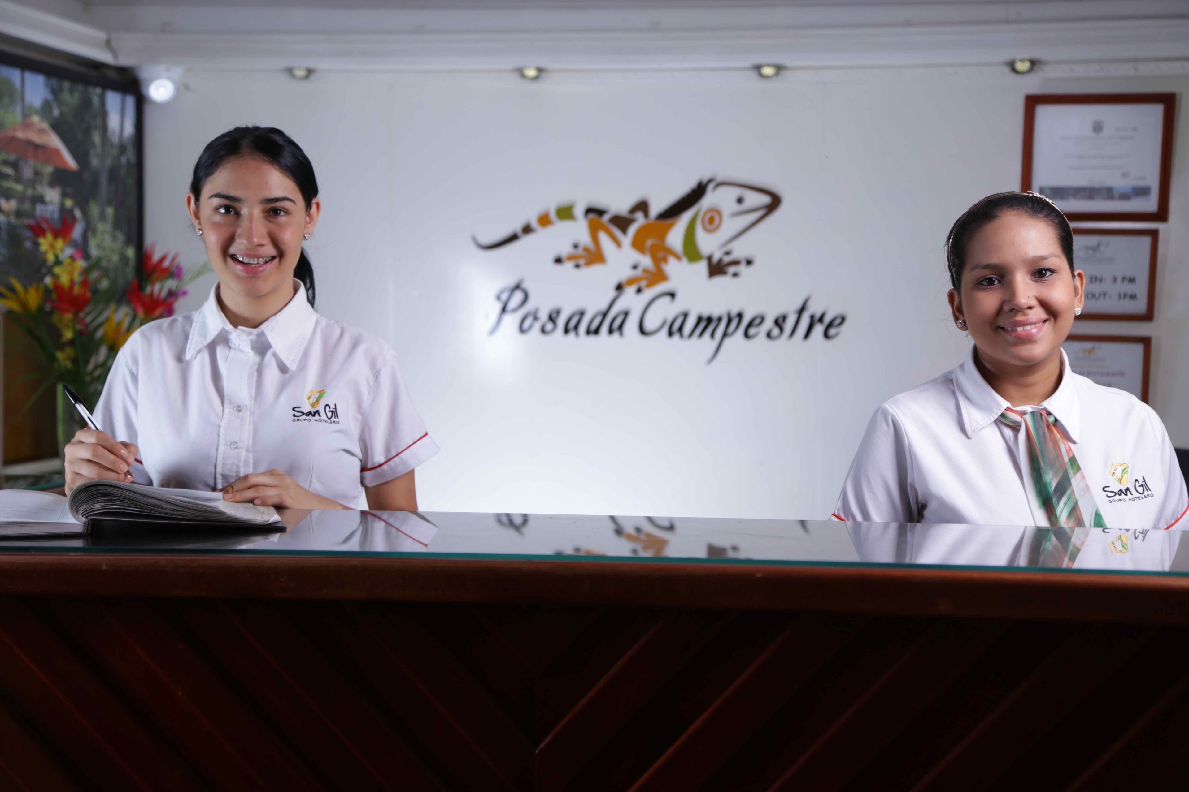 Dos recepcionistas en el mostrador de un hotel, con un logotipo y certificados enmarcados en la pared de fondo.