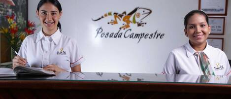 Dos recepcionistas en el mostrador de un hotel, con un logotipo y certificados enmarcados en la pared de fondo.