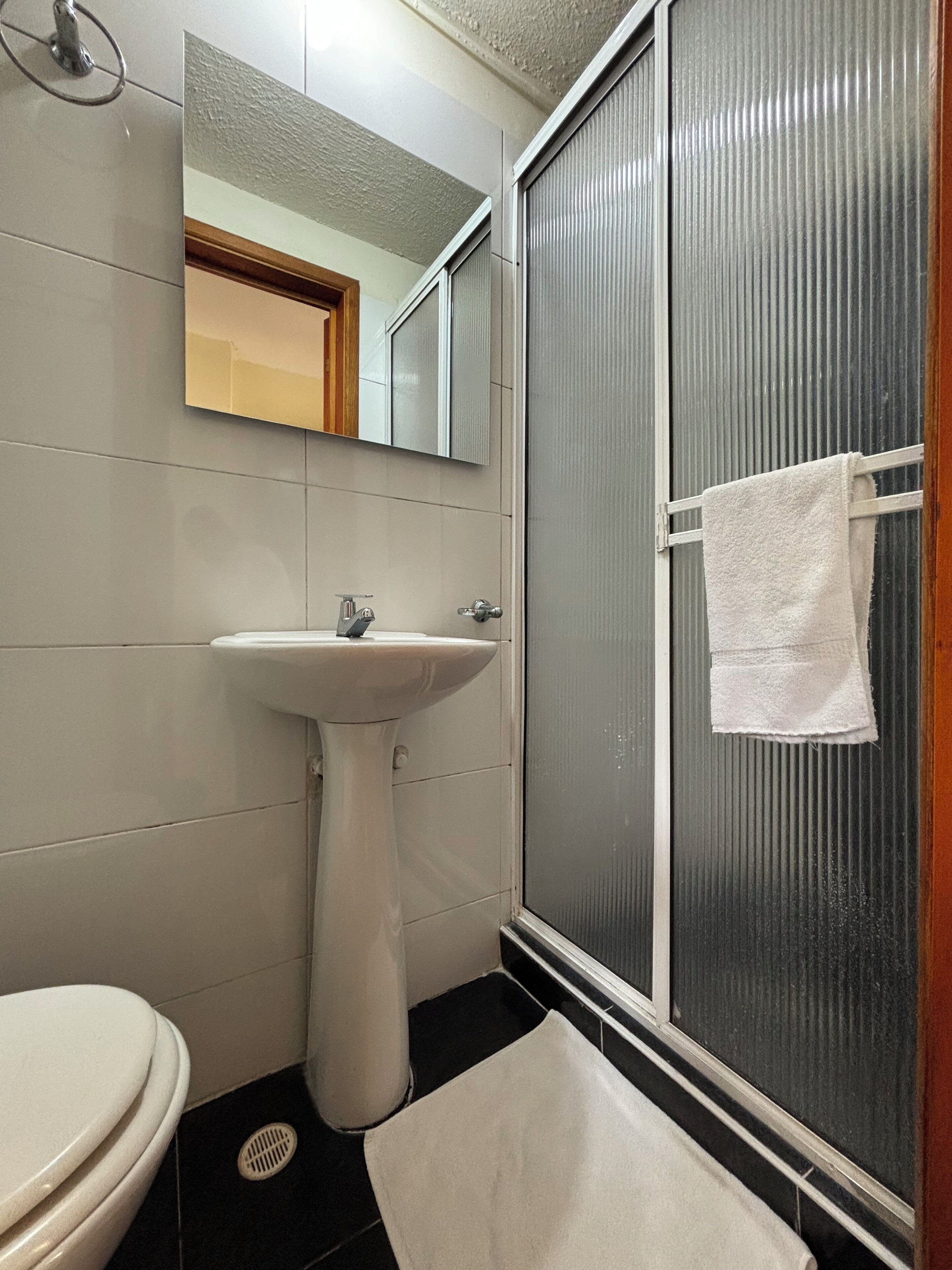 Un baño con lavabo pedestal, inodoro, espejo y ducha con una toalla colgada en la puerta.