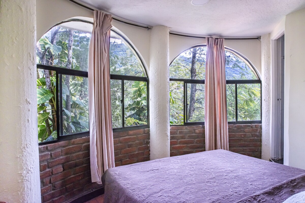 Una habitación con una cama, dos ventanas de arco con cortinas, una pared de ladrillo y vistas a la vegetación.