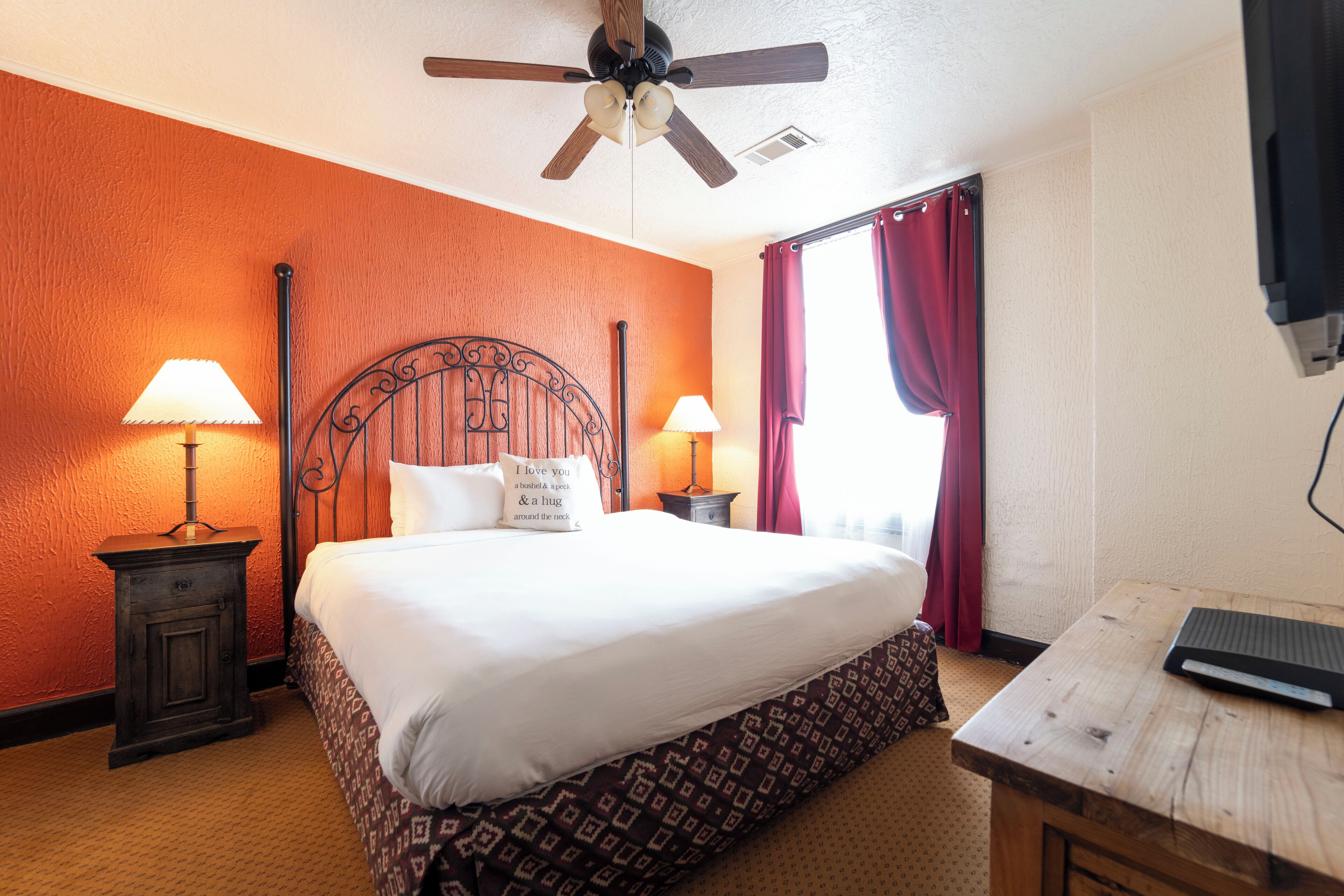 Una habitación de hotel con cama, dos lámparas de noche, una mesita de noche de madera, un escritorio, un ventilador de techo y una ventana con cortinas rojas.