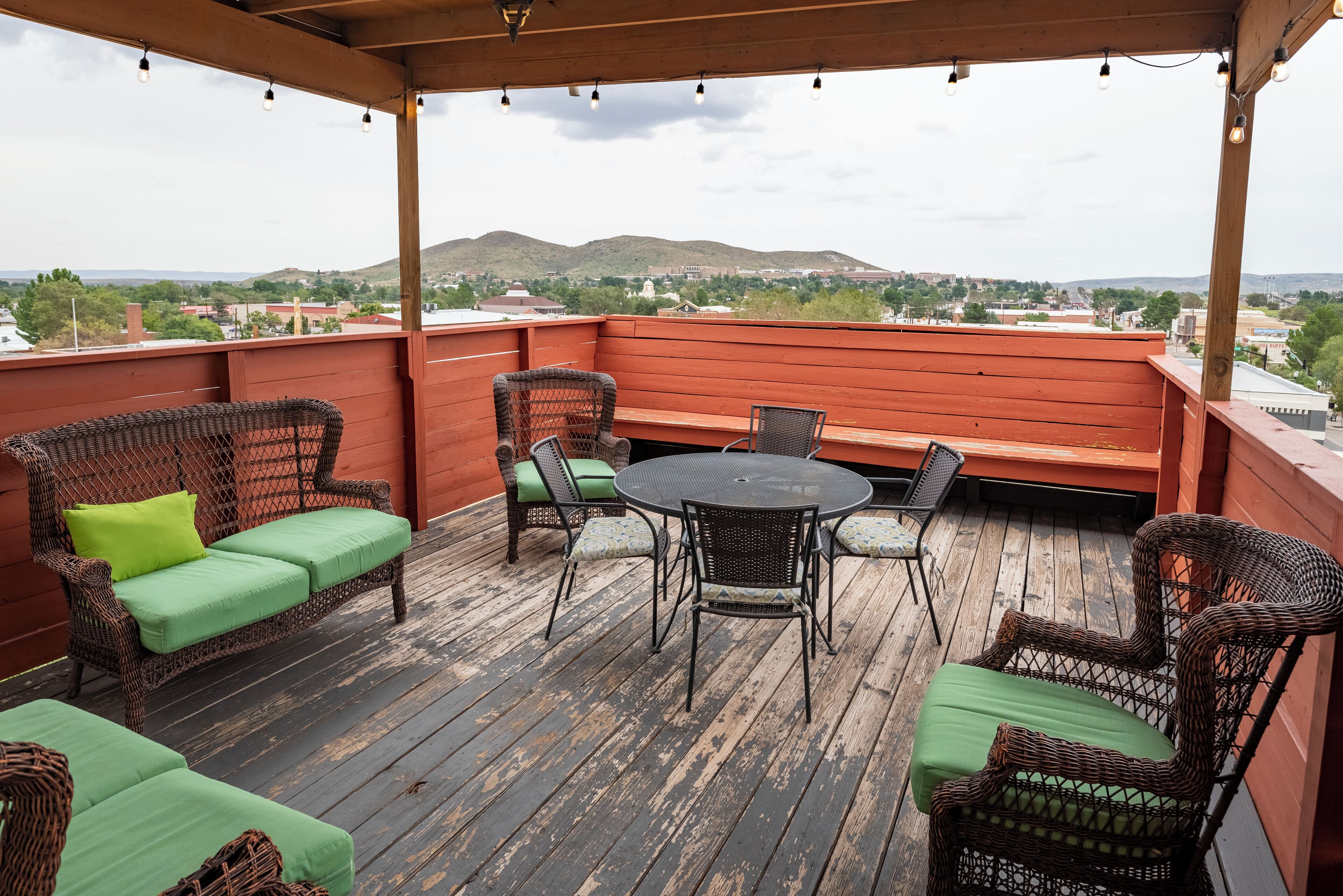 A covered outdoor seating area with wicker furniture and a round table, overlooking a cityscape and hills.