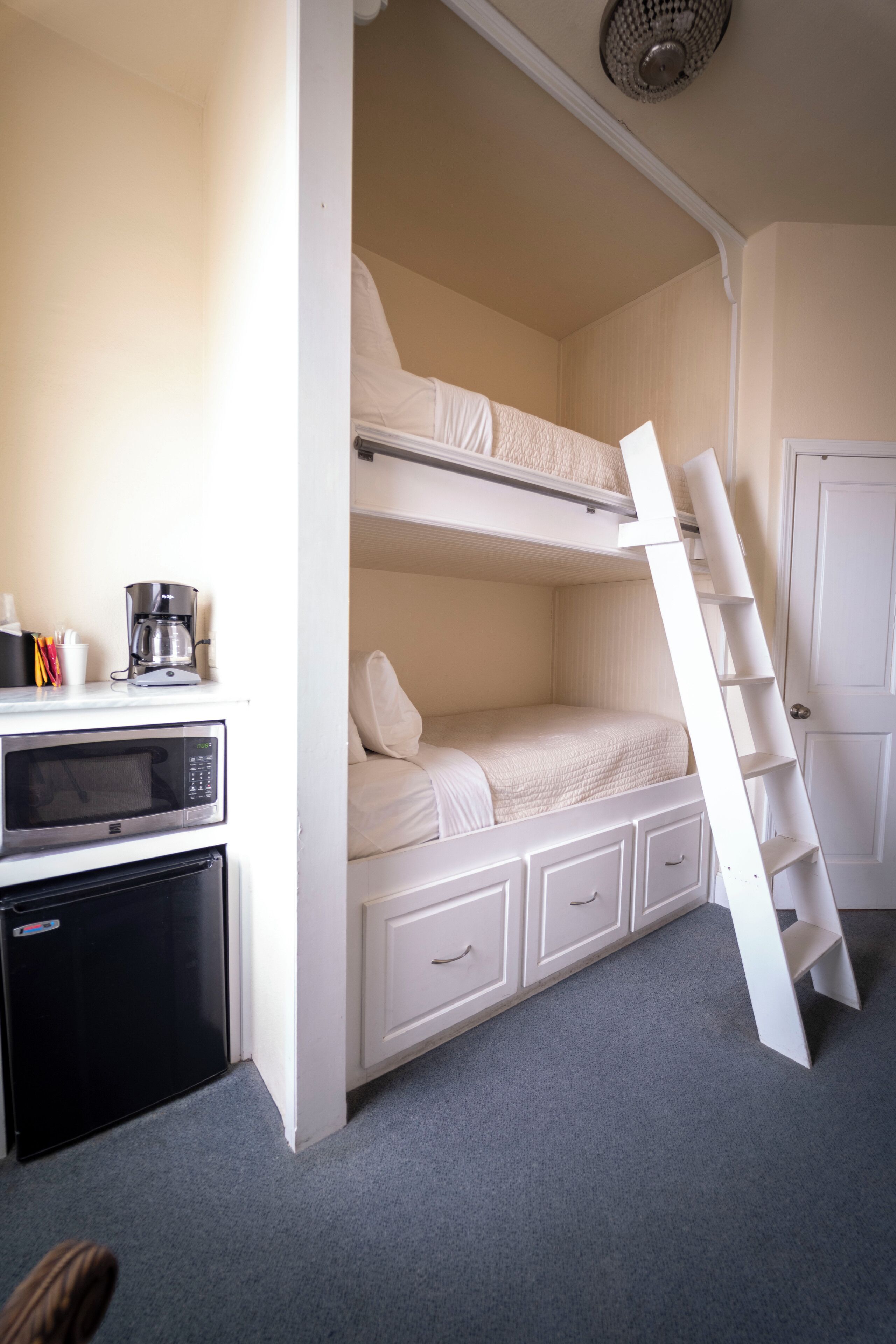 Een stapelbed met een ladder, een magnetron, een koffiezetapparaat en een koelkast in een kamer met een deur en een vloerbedekking.