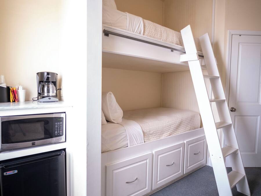 Een stapelbed met een ladder, een magnetron, een koffiezetapparaat en een koelkast in een kamer met een deur en een vloerbedekking.